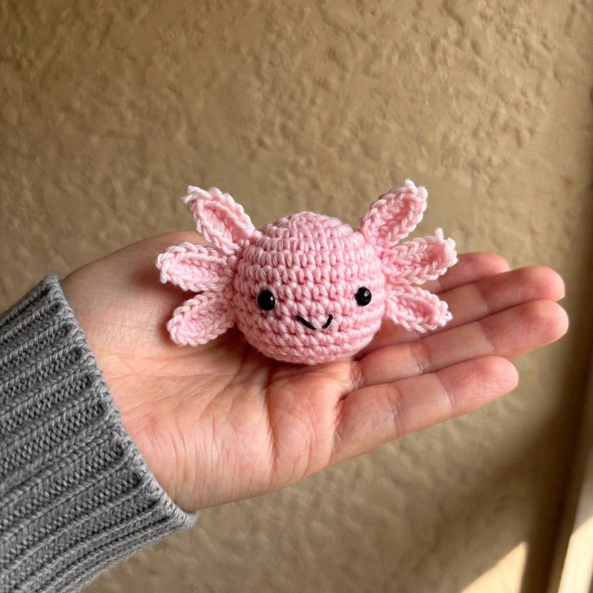 Pink crochet chubby axolotl amigurumi with frilly gills resting on an open hand