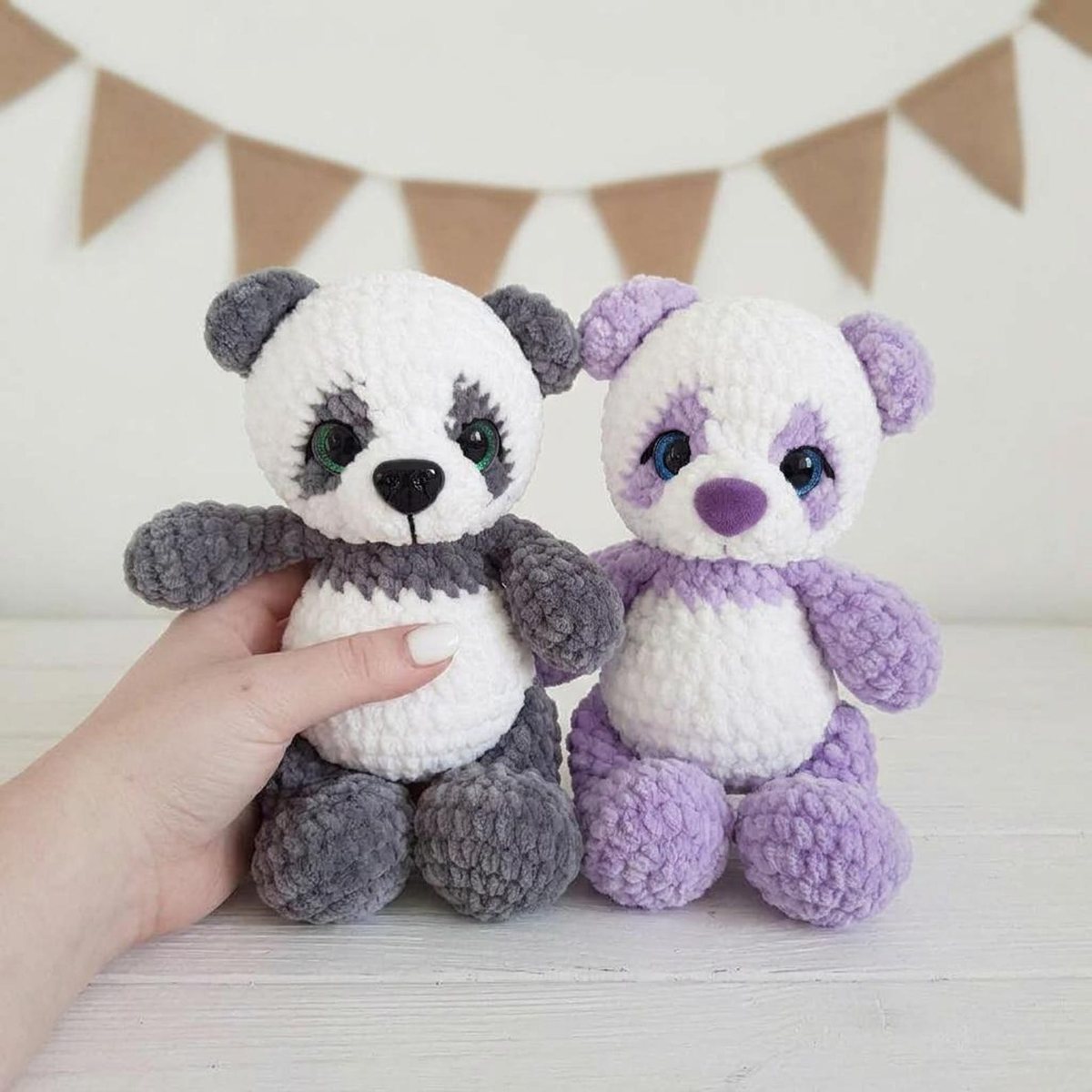 Two chenille crochet sitting pandas, one black and white and one lavender and white, held up in a pastel room with bunting
